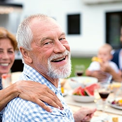 Ein älterer Mann sitzt lachend am gedeckten Pickniktisch mit einer Hand liebevoll auf seiner Schulter.