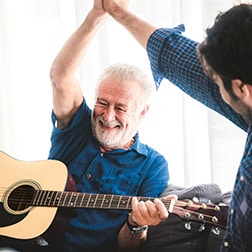 Ein Senior mit Gitarre hebt freudig die Hand zu einem High Five mit dem Pfleger.